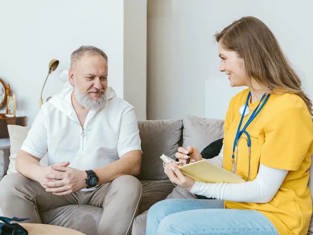 Eine Pflegekraft mit Stethoskop und ein älterer Mann lachen gemeinsam auf seinem Sofa.
